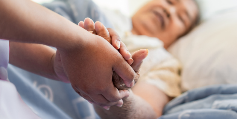 Fotografia de umas mãos de uma pessoa cuidadora a agarrar na mão de uma pessoa que está deitada numa cama.